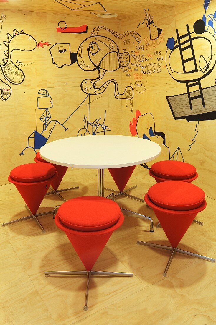 Modern, plywood paneled conference room with a white, round table and stools upholstered in orange fabric in front of a trendy wall decorated with cartoons (Red Bull Zentrale, Amsterdam)