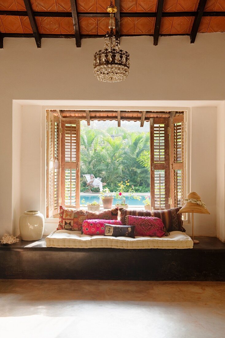 Floor cushions and scatter cushions on wooden platform below open window with shutters and view of pool in garden