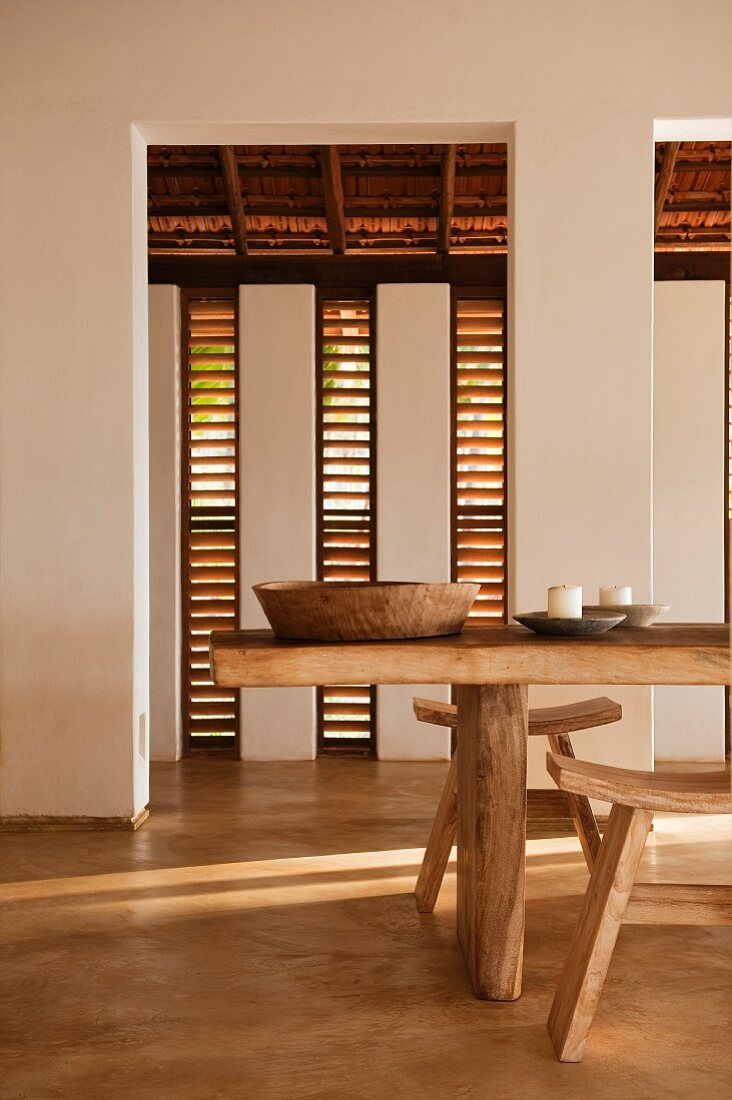Essbereich mit Lamellenfenstern und rustikalem Esstisch mit Hockern; auf dem Tisch eine einfache Holzschüssel