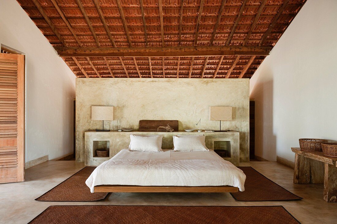 Geräumiges Schlafzimmer mit gemauertem Bett-Kopfende unter der Dachschräge; an der Wand ein rustikaler Wandtisch mit Körben