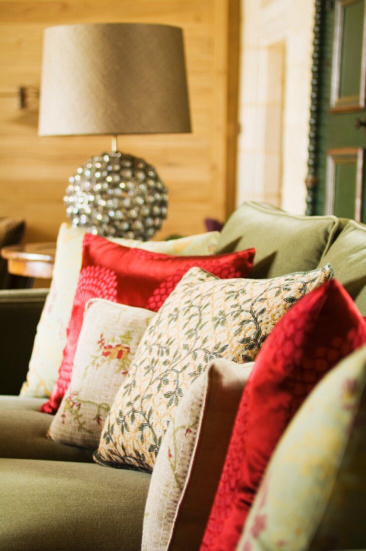 Gemusterte Kissen und teilweise mit rotem Satinbezug auf grünem Sofa neben Tischleuchte auf Beistelltisch