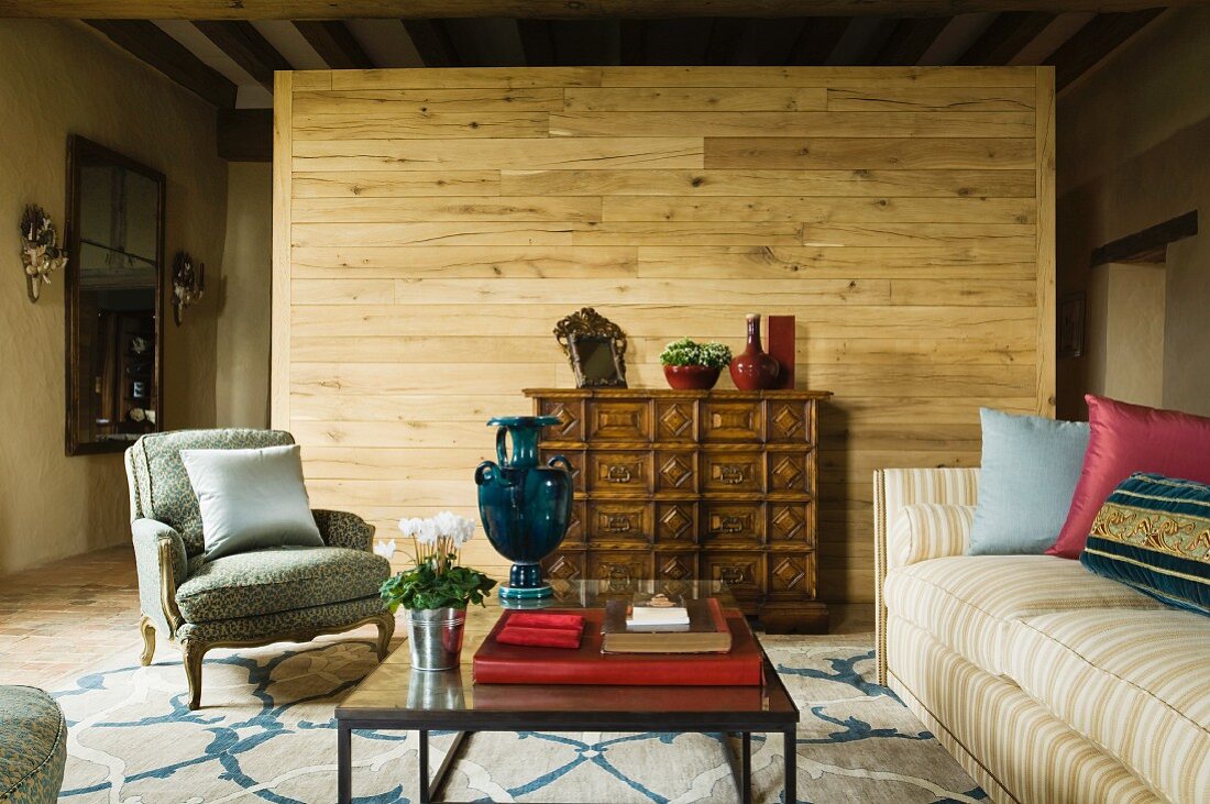 Traditioneller Sessel im Rokoko-Stil und Sofa um modernen Couchtisch vor Holz Raumteiler in rustikalem Wohnraum