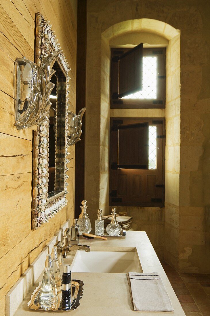 Verzierter Spiegel zwischen Wandleuchten an Holzwand über Waschtisch mit Parfumflakons neben Fensternische mit halbgeschlossenen Innenläden