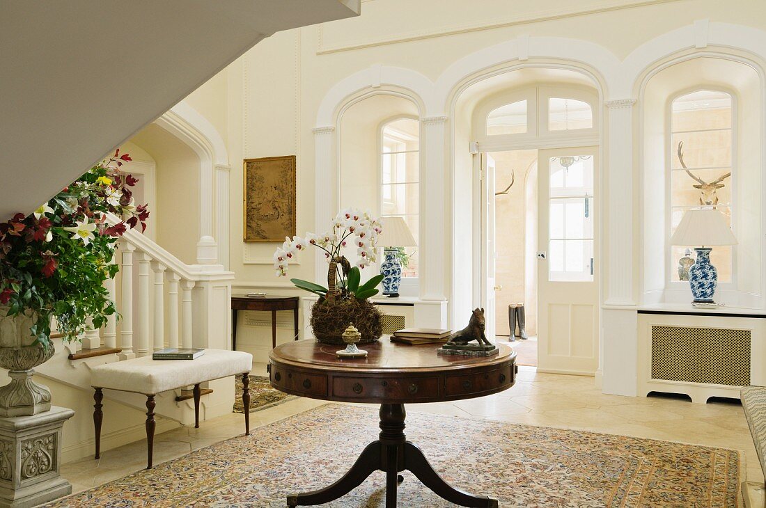 Round wooden table next to foot of staircase and arched doorway in foyer of English stately home
