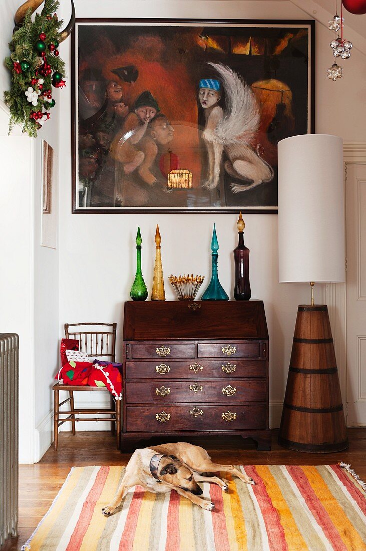 Festively decorated hallway with painting above coloured bottles on antique bureau; dog on striped rug