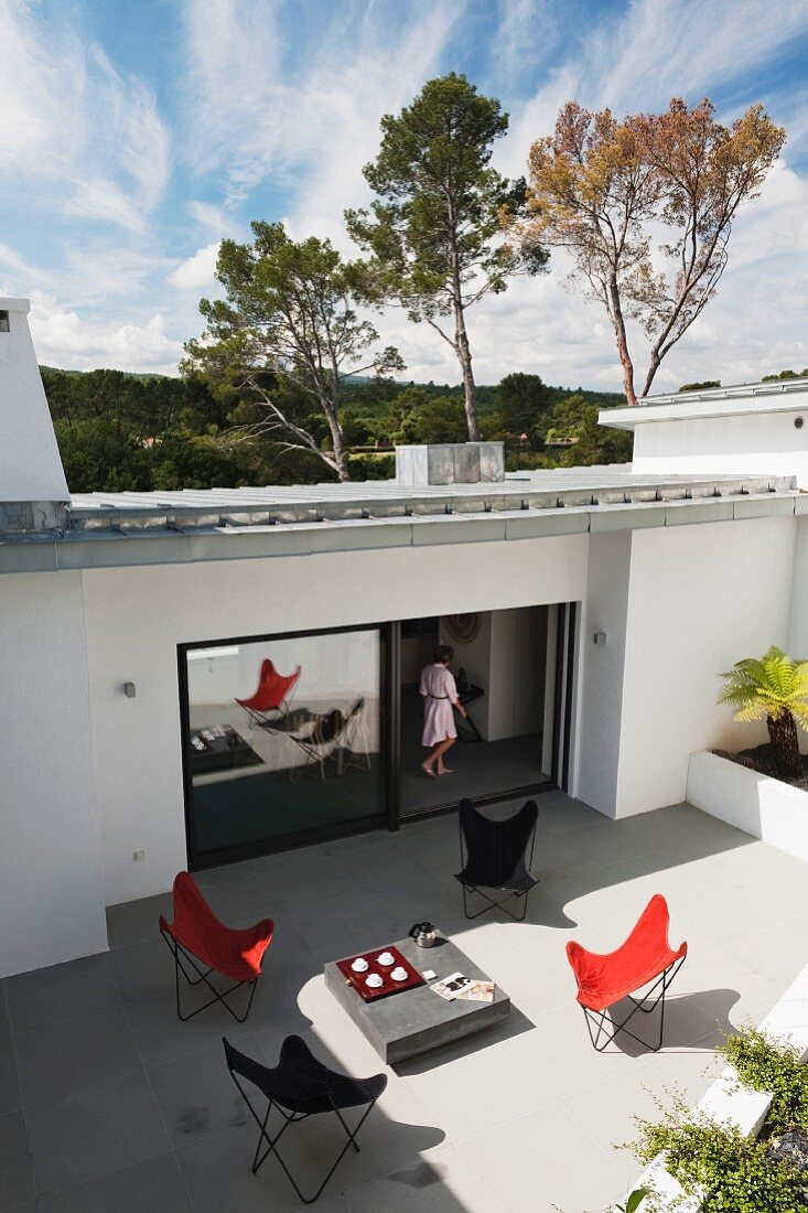 Blick auf grosse Terrasse eines modernen Wohnhauses mit verschiedenfarbigen Butterfly Sesseln und Frau in offener Terrassentür