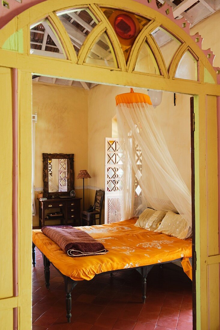 Blick durch offene Tür mit geschnitztem Rundbogen aus gelb lackiertem Holz auf Doppelbett mit gelber Bettwäsche
