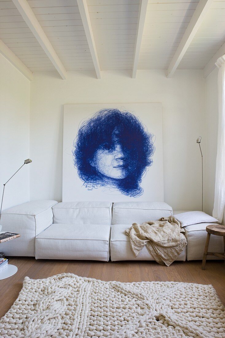 Wohnraum mit weißem Teppichvorleger und helle Polstercouch vor Wand mit Portraitzeichnung