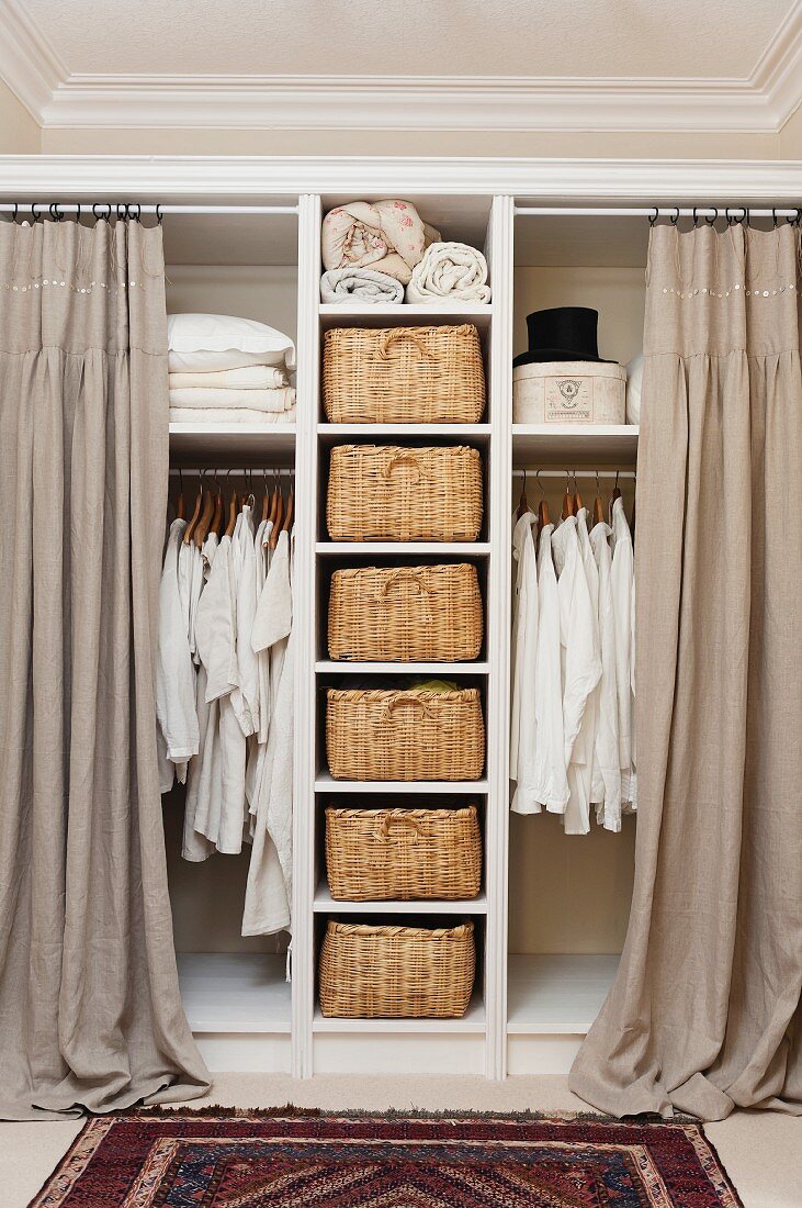 White shirts and linens hanging on rails in open storage with curtains. Wicker baskets form drawers on open shelving