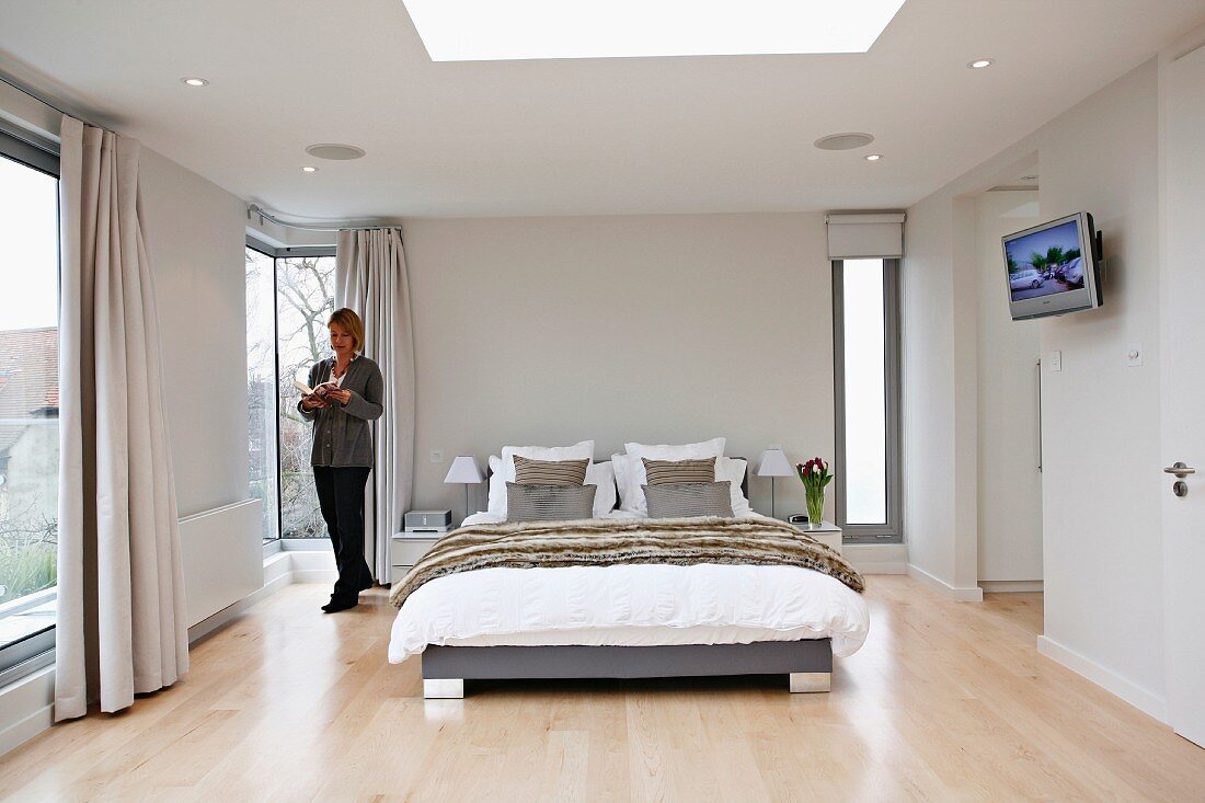 Frau steht neben Bett in elegantem Schlafzimmer mit raumhohen Fenstern