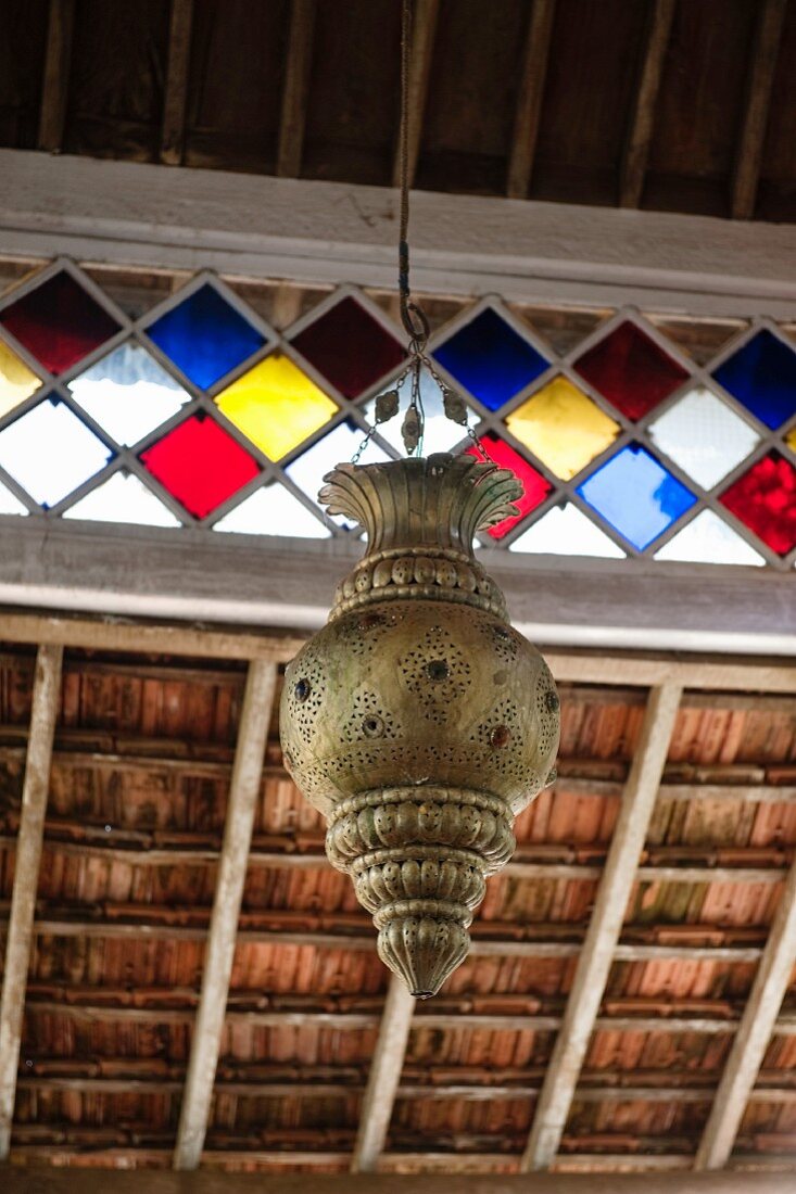 Indian-style metal pendant lamp hanging from wooden ceiling with stained glass ribbon window
