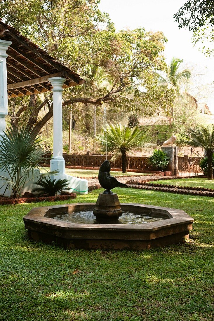 Brunnen mit Figur im Garten eines indischen Wohnhauses mit Vordach auf Säulen im Kolonialstil