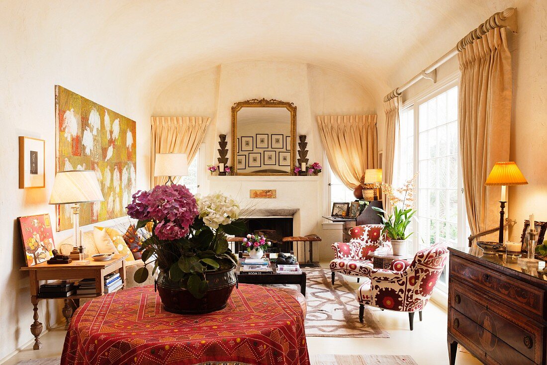 Sonniges Wohnzimmer mit offenem Kamin unter der Tonnendecke; im Vordergrund eine Vase mit violett und weiss blühenden Hortensien auf rundem Tisch