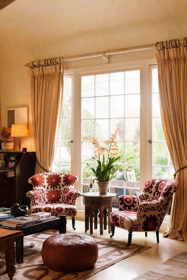 Wohnzimmer mit blumengemusterten Sesseln vor raumhohem Sprossenfenster; Im Vordergrund ein Polsterhocker aus braunem Leder