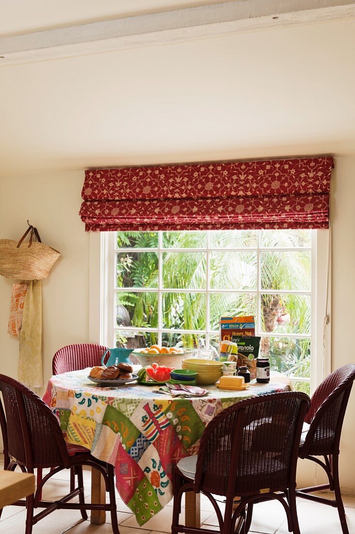 Runder Esstisch mit Patchwork-Tischdecke und Rattanstühlen vor großem Sprossenfenster mit gemustertem Stoffrollo