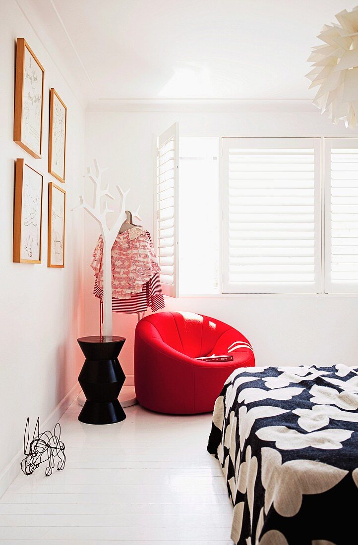 weiße Schlafzimmerecke mit rotem Polstersessel neben stilisiertem Baum als Kleiderständer vor Fenster mit weissen Innenläden