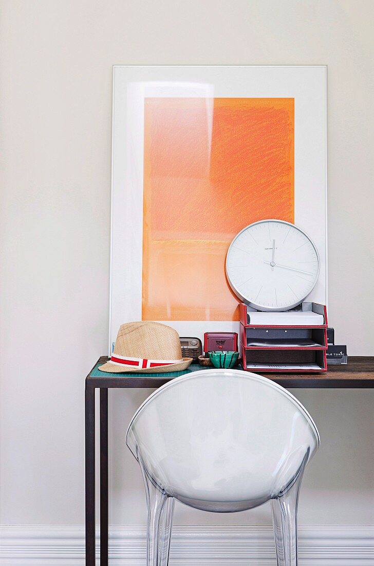 Schalenstuhl aus weißem Kunststoff vor Arbeitstisch und modernes Bild an Wand gelehnt