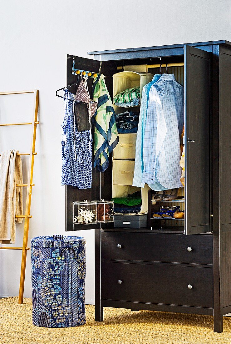 Clothing hanging in open, black wardrobe with drawers next to patterned waste-paper basket