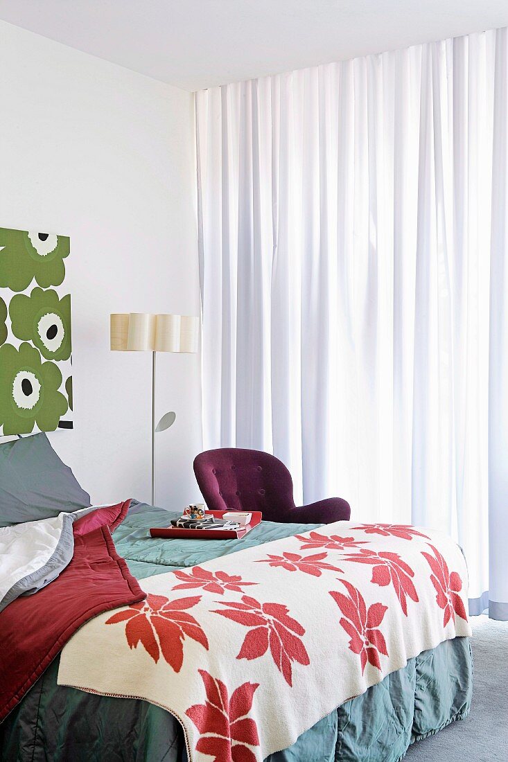 Bett mit zweifarbiger Tagesdecke und stilisierte Blumenlampe in Schlafzimmerecke mit großformatigen floralen Mustern