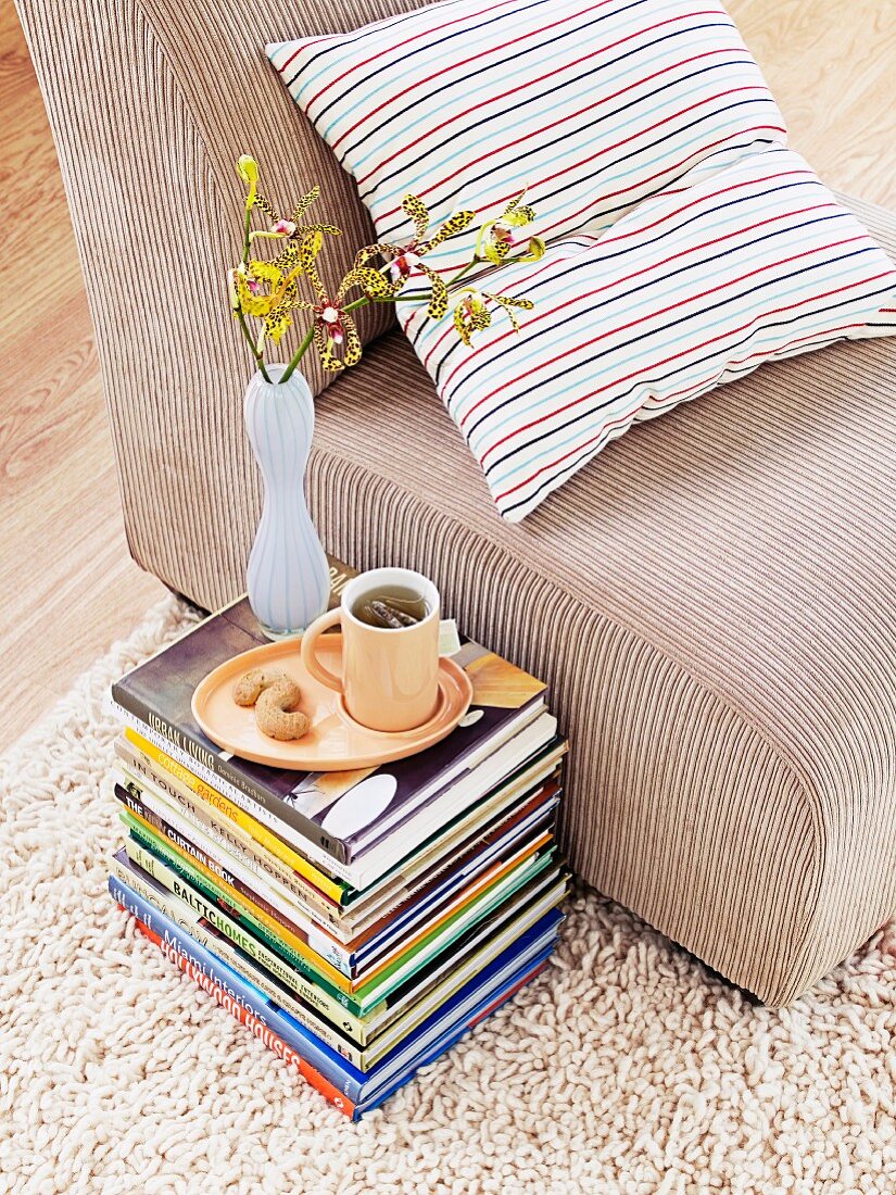 Mug of tea and vase of flowers on stack of books on floor next to armchair with beige corduroy cover and striped cushion