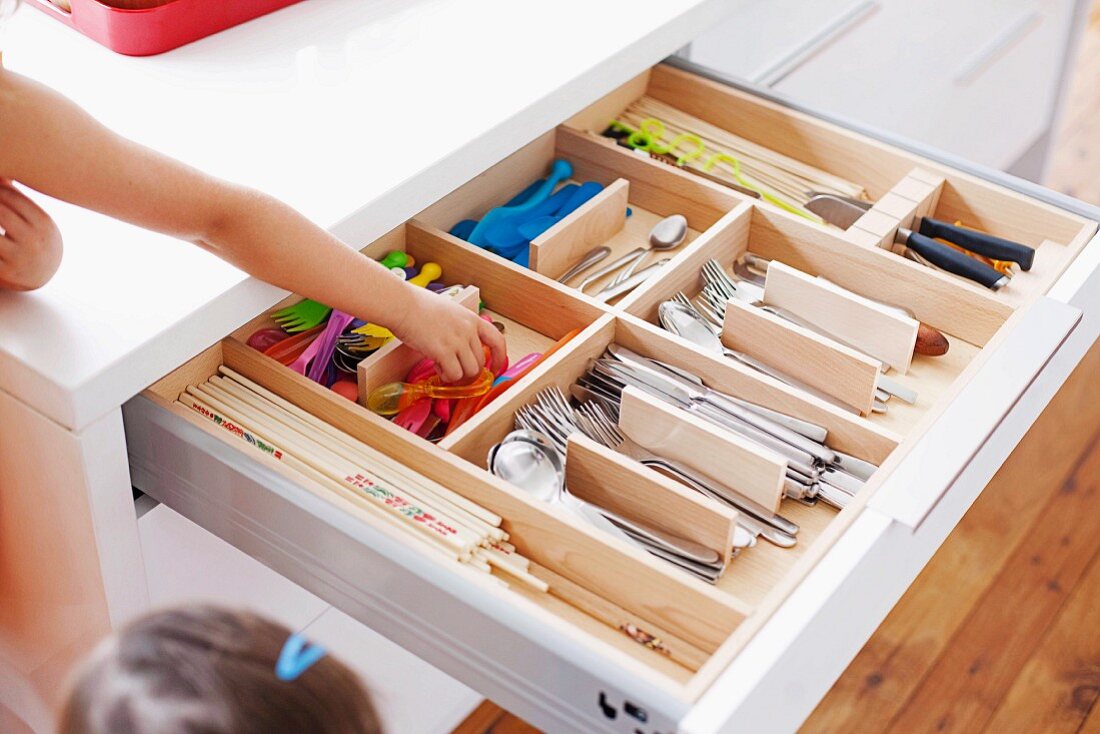 Open cutlery drawer with various cutlery in wooden insert and child's hand reaching for child's cutlery