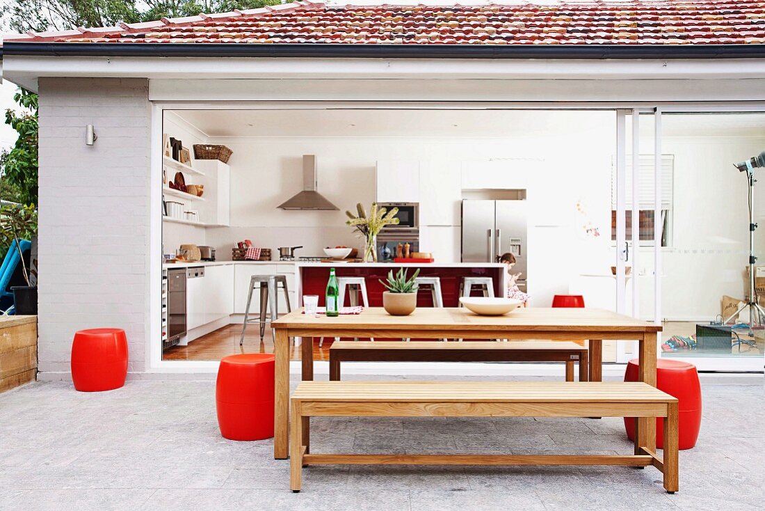 Furnished terrace area with red, barrel-shaped stools in front of open glass wall; complete view into mainly white kitchen-dining area of bungalow