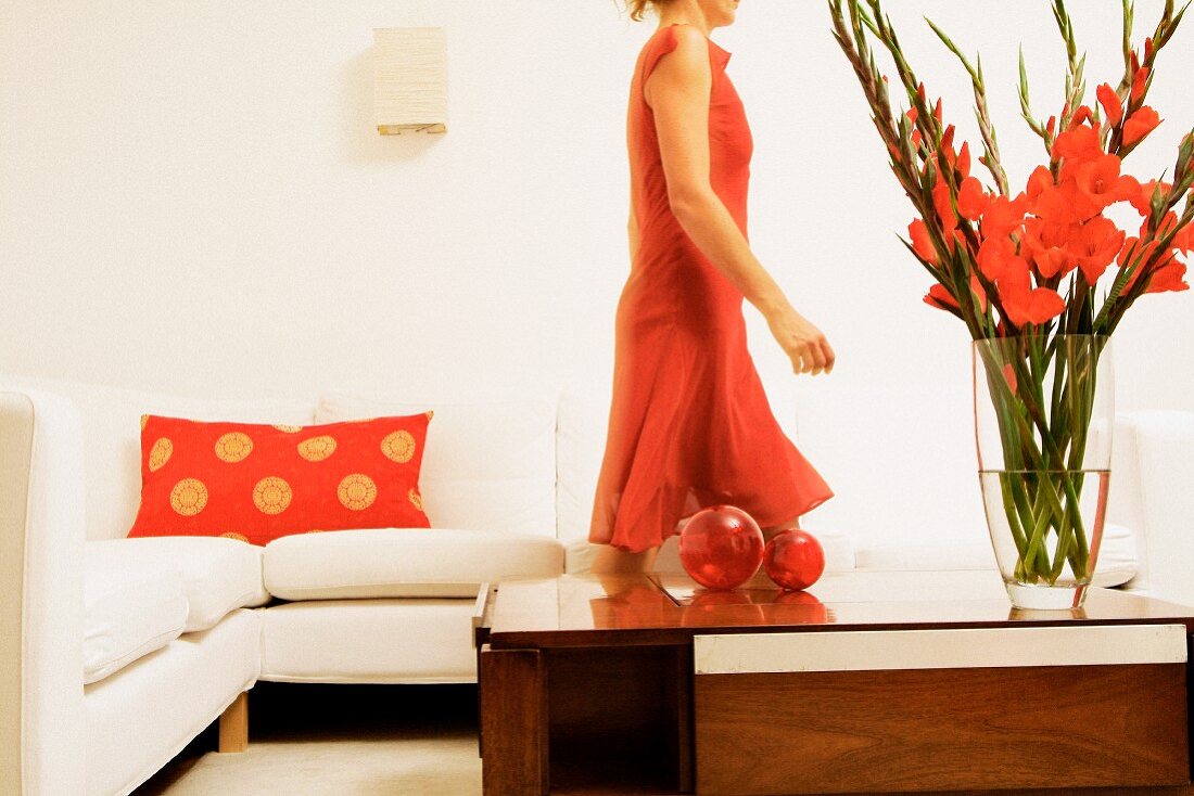 Wohnzimmertisch mit roten Gladiolen, dahinter Frau im roten Kleid vor weißem Sofa