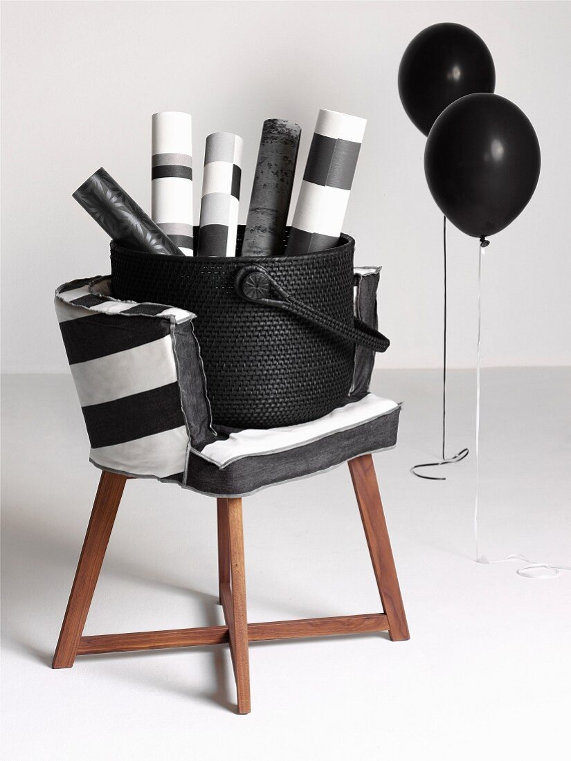 Rolls of paper in basket on chair with wooden frame and black and white cover next to black balloons on metal frames