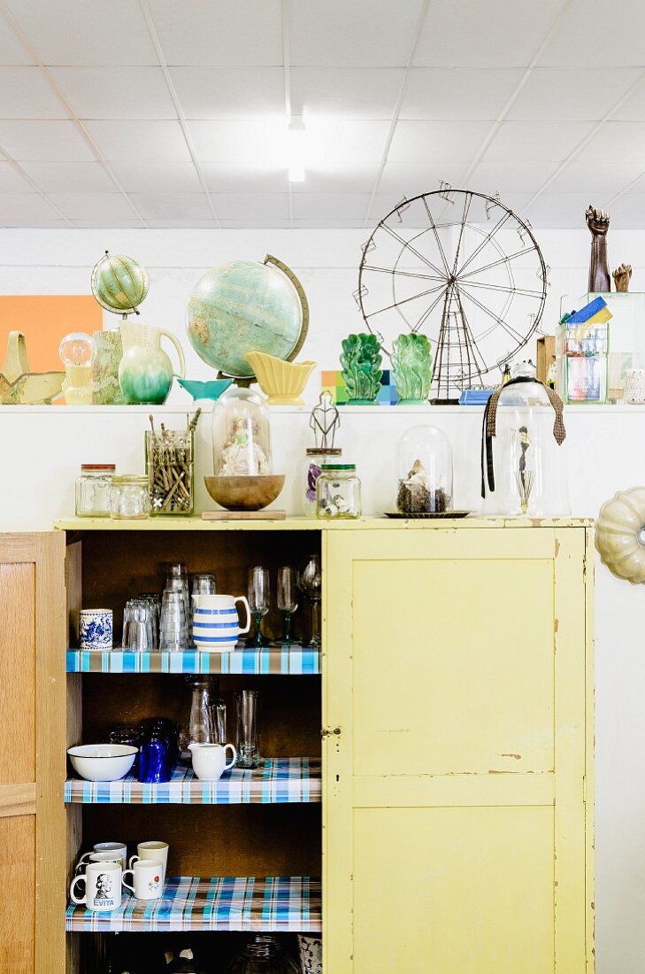 Simple cupboard with open door and view of crockery inside against partition with collectors' items on top