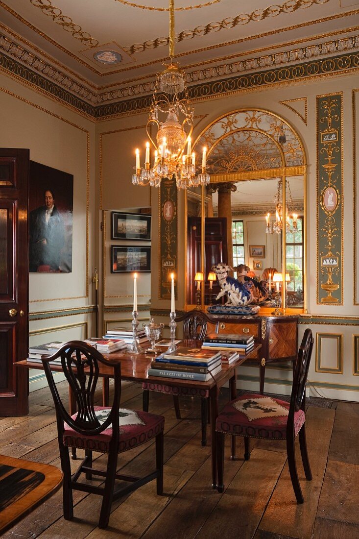 Lit candles in chandelier above dining table and upholstered chairs; custom-made replica mirror surrounded by gilt wall panelling in background