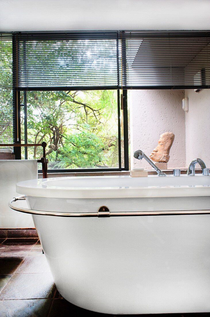 Freistehende Retro Badewanne mit Handtuchhalter vor Fenster mit halb geschlossener Jalousie und Blick in Garten