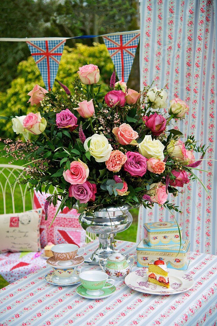 Romantisch gedeckter Tisch im Garten mit prallem Rosenstrauß in silbernem Kelch zu englischem Teeservice und Tischdecke mit Blümchendessin