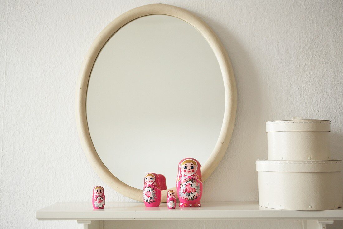 Rosa Matrjoschka-Puppen und weiße Schachteln vor ovalem Spiegel auf einem Wandboard