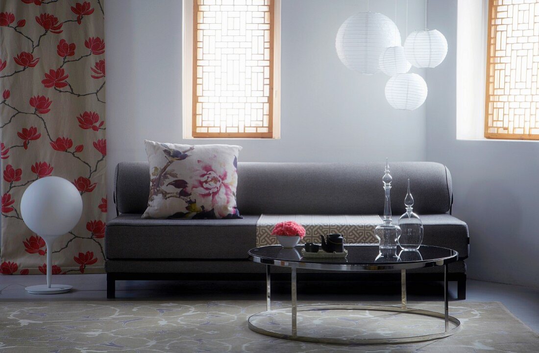 Grey sofa with floral scatter cushion in front of pendant paper lamps; glass carafes on shiny coffee table in foreground