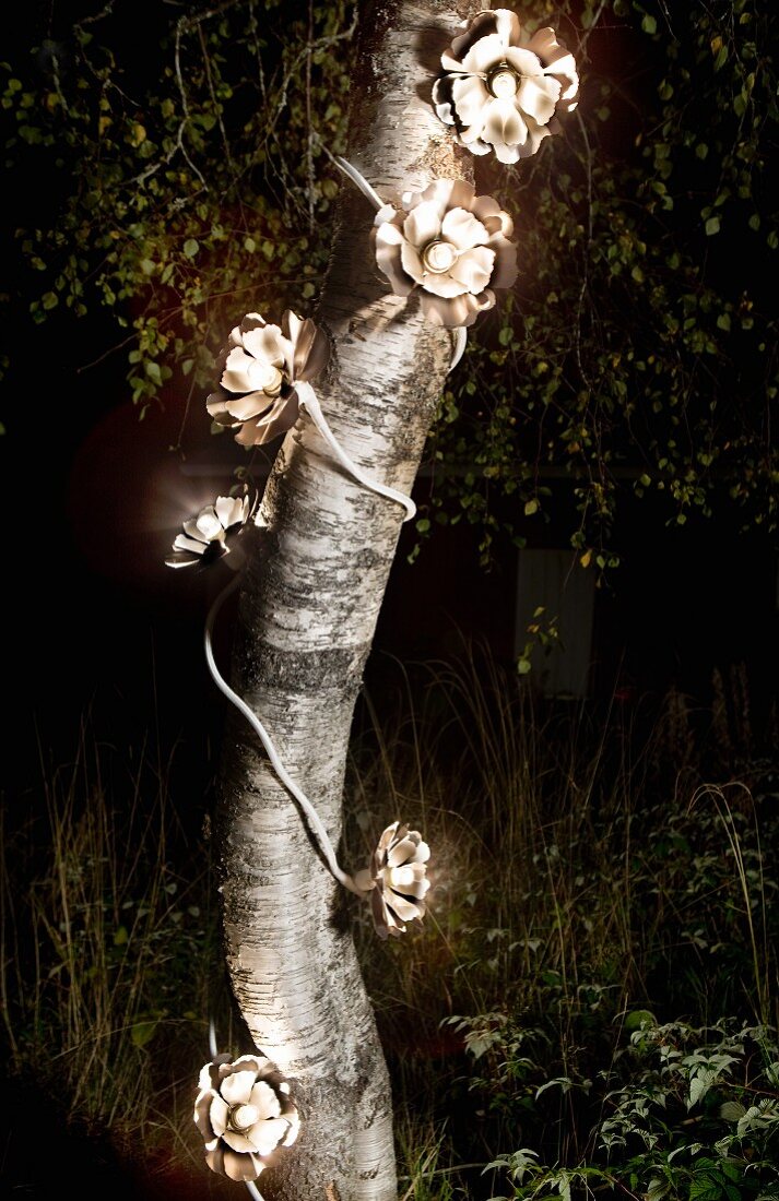 Lichterkette mit Blütenmotiv ist um Baumstamm gewickelt und erstrahlt im nächtlichen Wald
