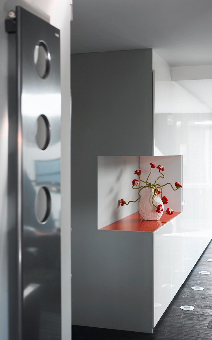 White, glossy, fitted cupboards with cut-out corner and orange shelf holding crocheted, floral ornament