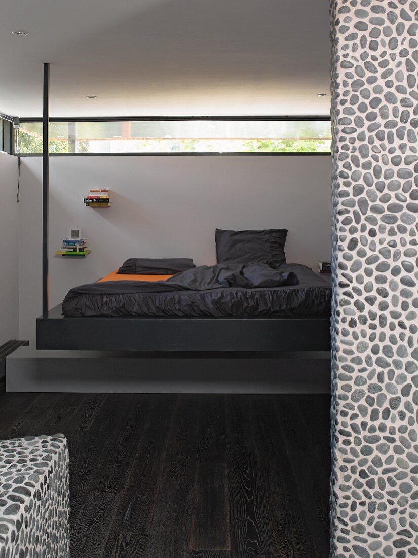 Designer bed suspended from ceiling by metal poles below circumferential ribbon window in bedroom with dark parquet floor; pebble mosaic tiled wall in foreground