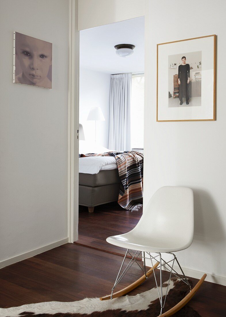View into bedroom with upholstered bed; white, modern, designer rocking chair on animal-skin rug and photos on white walls in anteroom