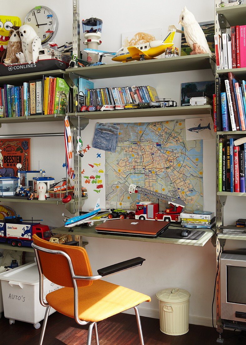 Volles Regal im Jungen-Kinderzimmer mit vielen Büchern und diversen Spielsachen, am Arbeitsplatz mit Holzstuhl liegt ein Laptop und dahinter ist eine Landkarte an der Wand