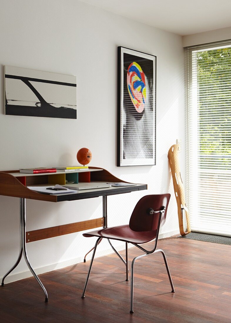 Modern desk and chair below abstract pictures on white wall; wooden sculpture leaning on wall in front of French window with blinds in background