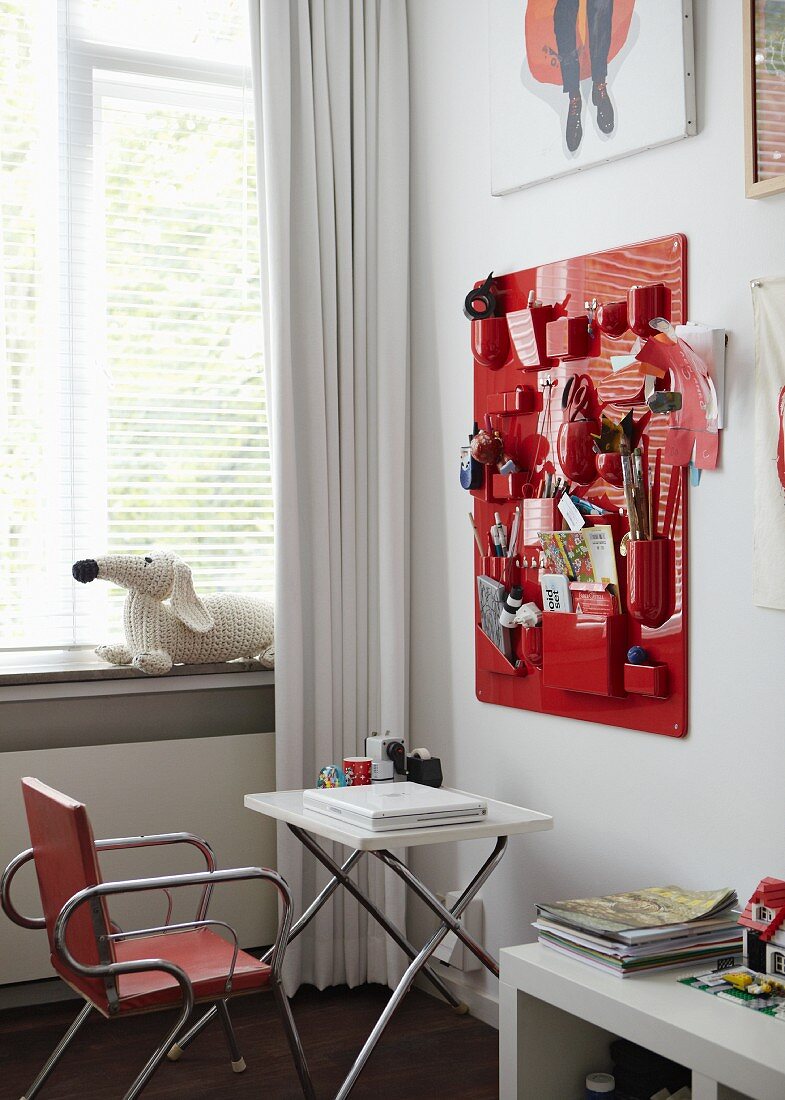 Mädchenhaftes Jugendzimmer mit roter Pinwand zur Aufbewahrung diverser Utensilien, davor ein weißer Klapptisch mit Laptop und einem Metallpolsterstuhl, vor dem Fenster ein weißer Vorhang