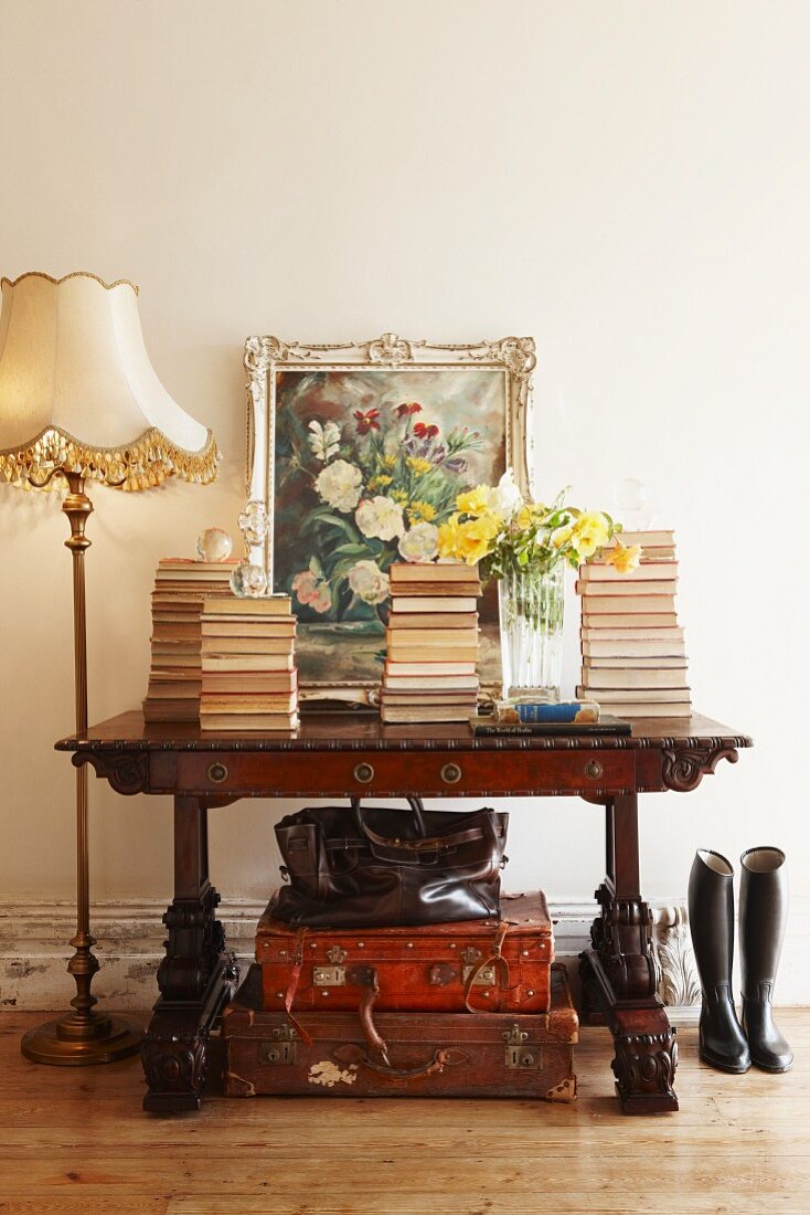 Antiker Schreibtisch mit Bücherstapeln und angelehntem Ölgemälde, darunter Lederkoffer, eine Biedermeier Stehlampe steht seitlich