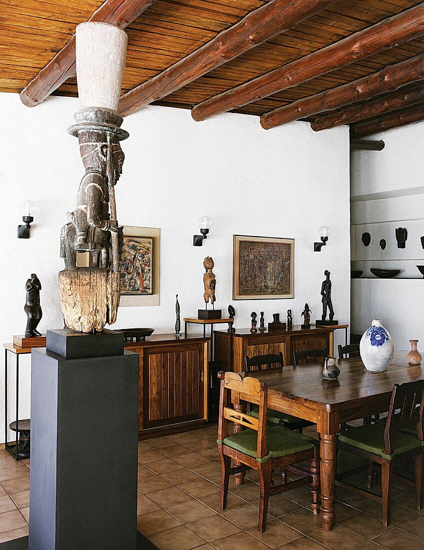Afrikanische Skulpturen teilweise auf Stele und auf Sideboard im Esszimmer mit Holzbalkendecke und modernem Flair