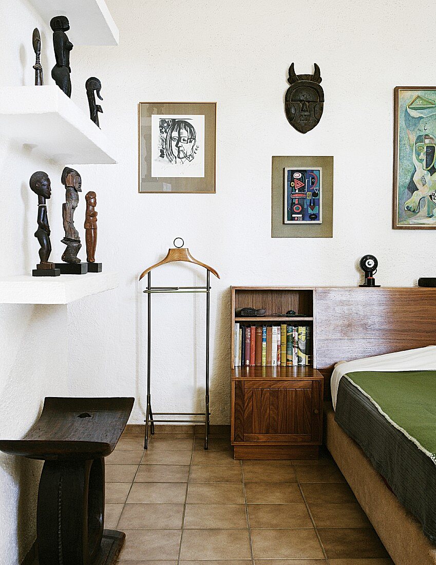 Holzhocker unter Regal mit afrikanischen Skulpturen gegenüber teilweise sichtbarem Bett in Schlafzimmerecke