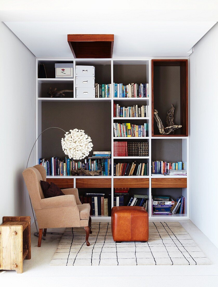 Smart fitted shelving in reading corner with traditional wing-backed chair and foot stool combined with pretty designer lamp