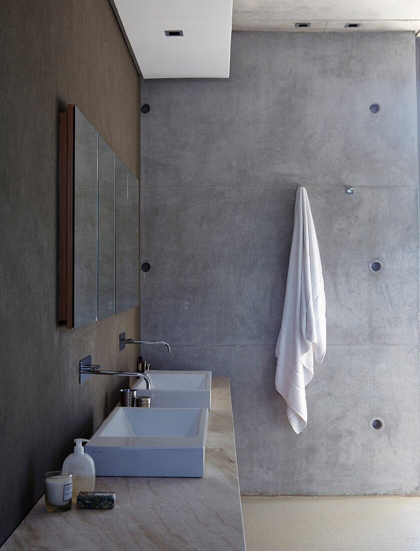 Stone washstand with counter-top basin and wall-mounted tap fittings on dark painted wall; towel on hook on purist exposed concrete wall