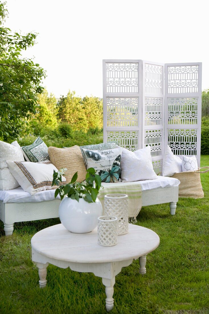 Vasen auf weißem Beistelltisch vor Sitzbank mit Kissen und Paravent im Garten