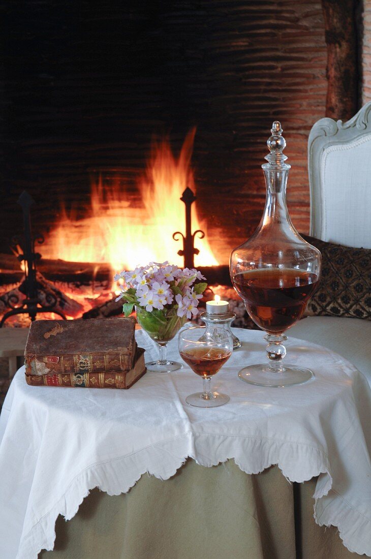 Nostalgisch gedeckter, runder Tisch mit Portweinkaraffe und Weinglas vor brennendem Kaminfeuer