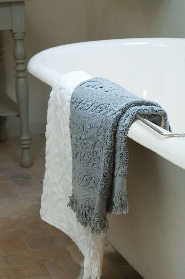 Graues und weisses Handtuch über dem Badewannenrand einer freistehenden Badewanne hängend