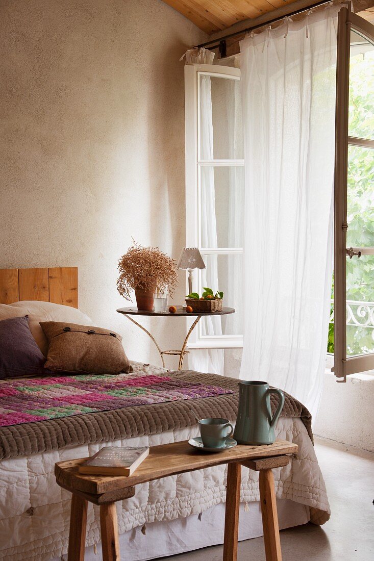 Holzbank vor Bett mit Tagesdecke; seitlich duftiger Vorhang im offenen Fenster